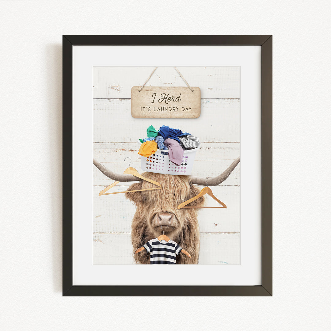 A framed image depicting a cow with a basket of laundry on its head, accompanied by a sign that reads "I herd, it's laundry day".