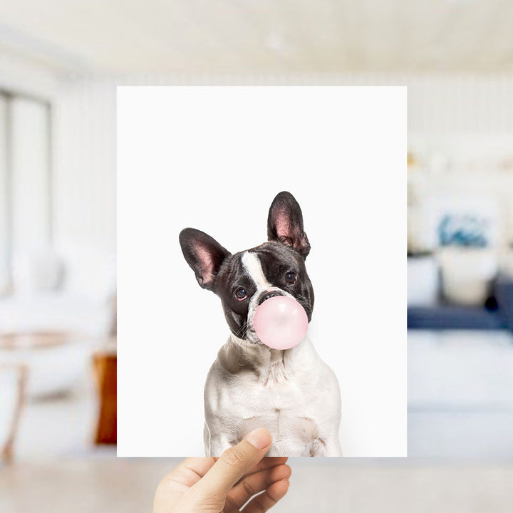 A hand is holding up a picture of a dog blowing a bubble with a pink bubble gum bubble.