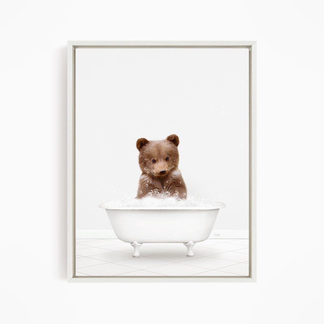 A cute brown bear sitting in a white bathtub filled with bubbles, looking directly at the camera.