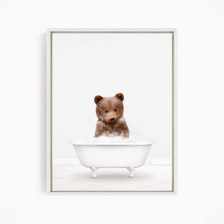 A cute brown bear sitting in a white bathtub filled with bubbles, looking directly at the camera.