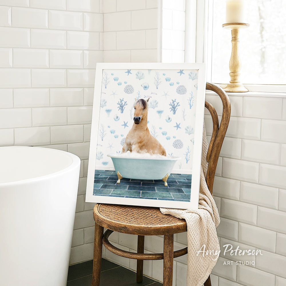 A framed image of a horse sitting in a bathtub, surrounded by blue and white seashells and starfish, is displayed on a wooden chair in a bathroom setting.