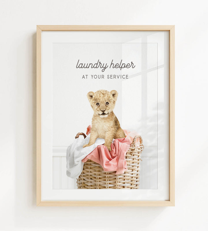 A framed image of a young lion cub sitting in a wicker laundry basket, with a pink blanket and white cloth inside.