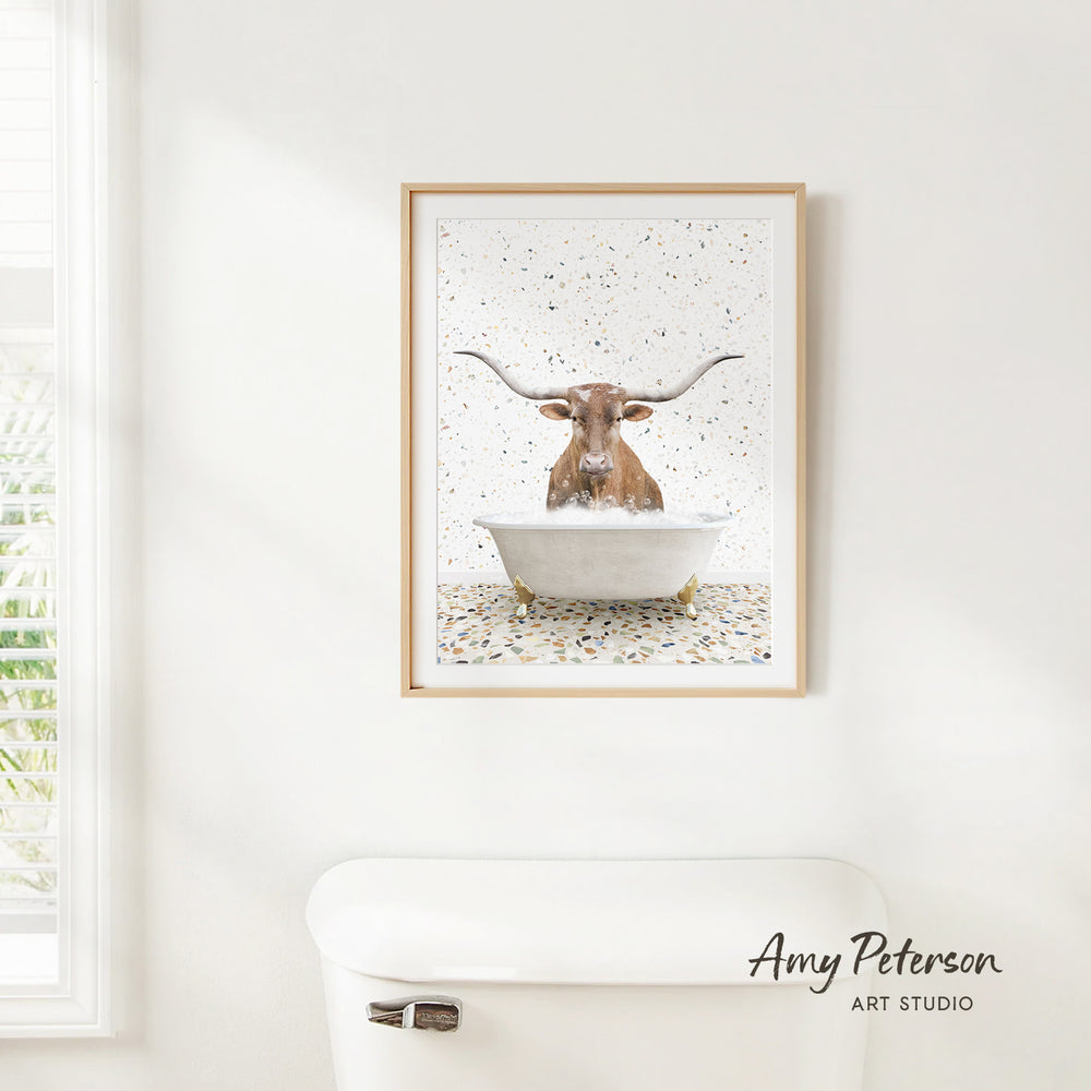 A framed print of a bull with horns sitting in a bathtub is hanging on a white wall.