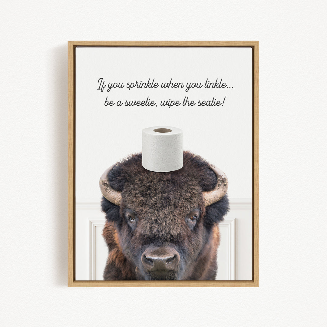 A framed image of a bison with a roll of toilet paper on its head, accompanied by a humorous text that reads "If you sprinkle when you tinkle... be a sweetie, wipe the seattle!"