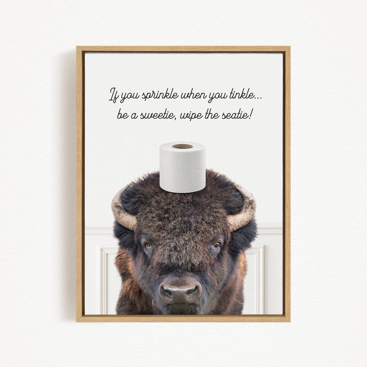 A framed image of a bison with a roll of toilet paper on its head, accompanied by a humorous text that reads "If you sprinkle when you tinkle... be a sweetie, wipe the seattle!"