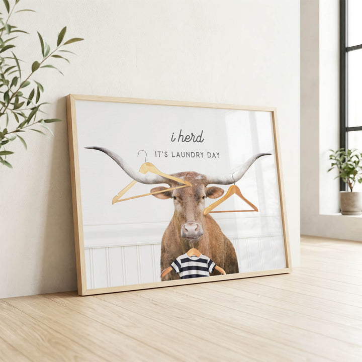 A framed poster with a bull's head and a person wearing a striped shirt, along with the text "It's laundry day" and the phrase "i herds" written above the bull's head.
