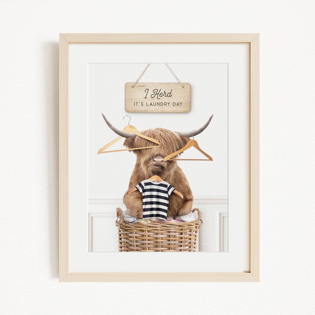 A framed image depicting a large, furry animal, possibly a yak or a yak dog, sitting in a wicker basket and holding a sign that says "I herd it's laundry day".