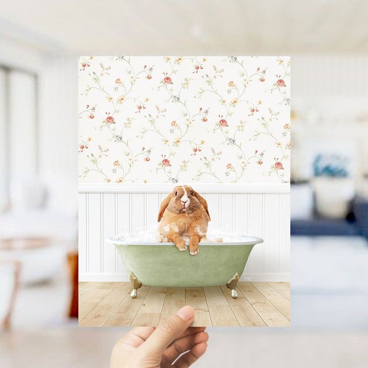 A hand holds up a photograph of a brown rabbit sitting in a green bathtub filled with bubbles, with a floral wallpaper in the background.