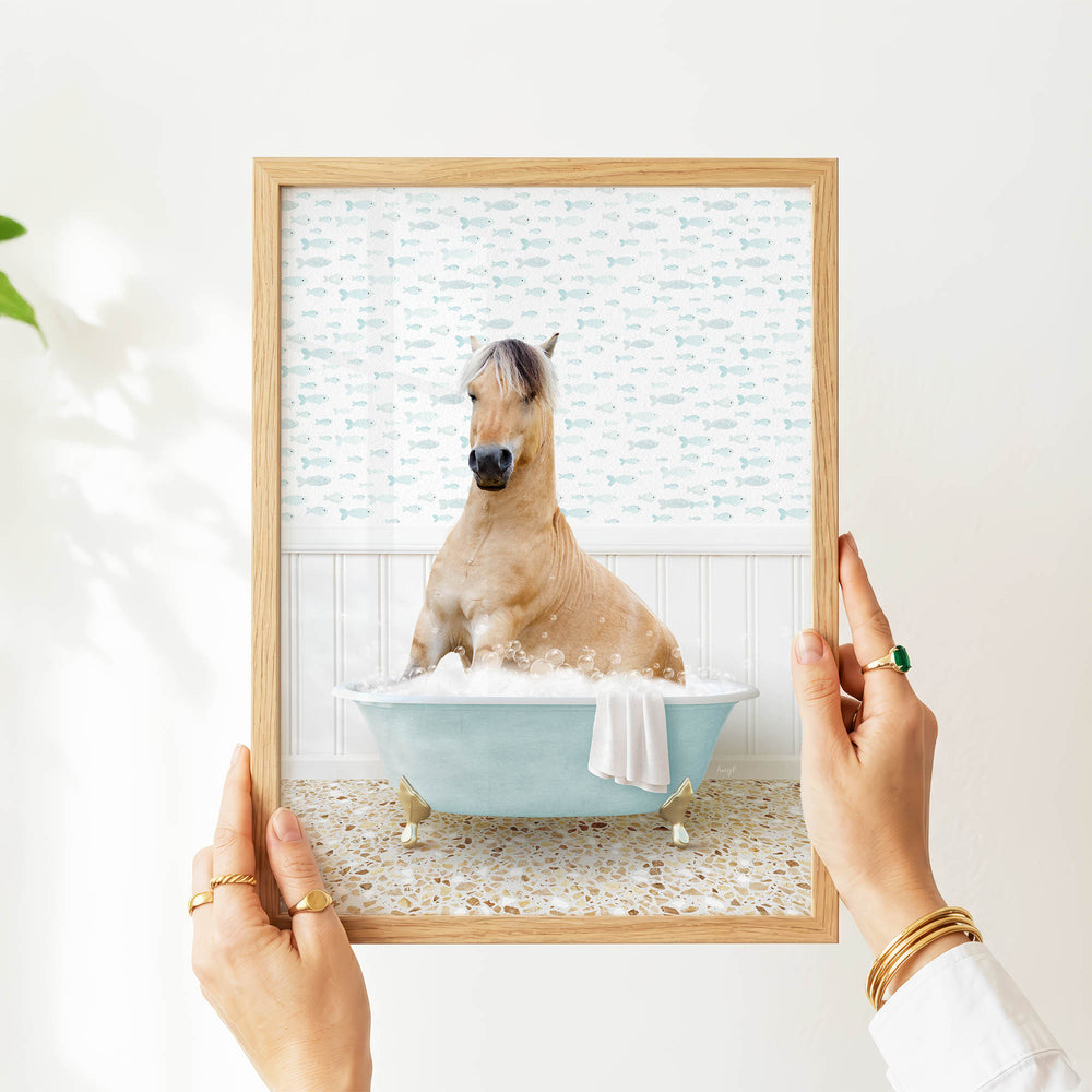 A person's hands are holding a framed picture of a horse sitting in a bathtub filled with bubbles, with a patterned wall behind it.