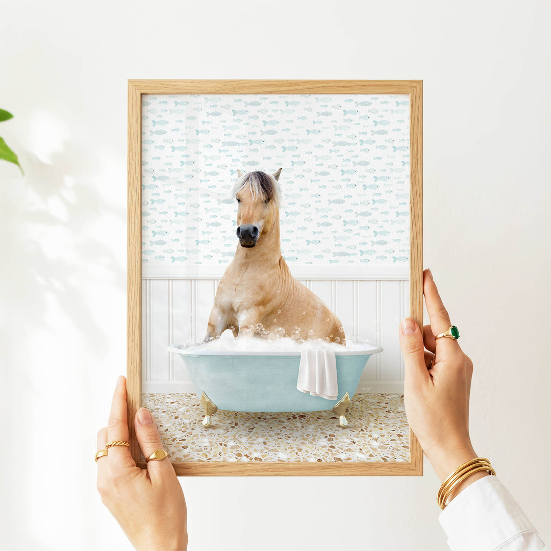A person's hands are holding a framed picture of a horse sitting in a bathtub filled with bubbles, with a patterned wall behind it.