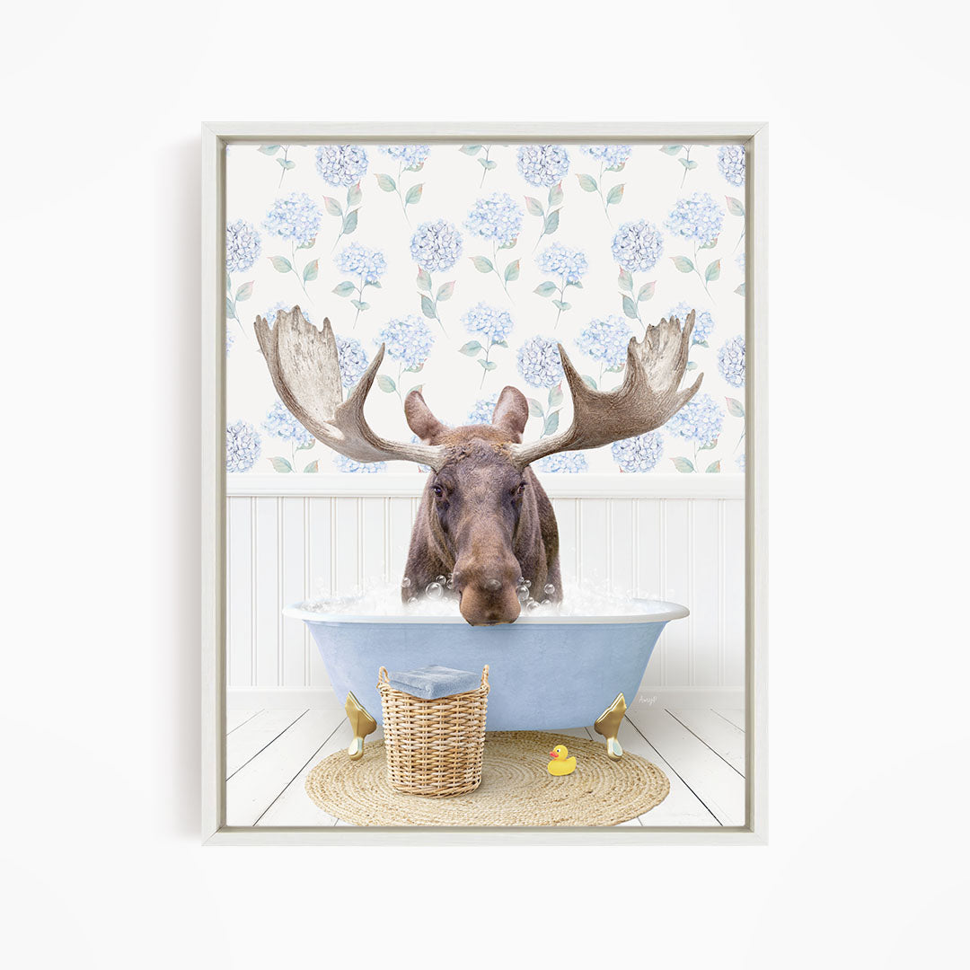 A moose with large antlers is sitting in a blue bathtub filled with water, surrounded by a wicker basket and yellow rubber ducks.