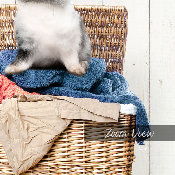 A cat sitting on top of a wicker basket filled with clothes, including a blue towel and a red towel.