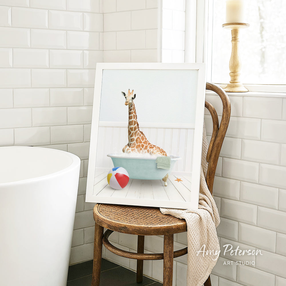 A framed image of a giraffe sitting in a bathtub, surrounded by a beach ball and a towel, is displayed on a wooden chair in a bathroom setting.