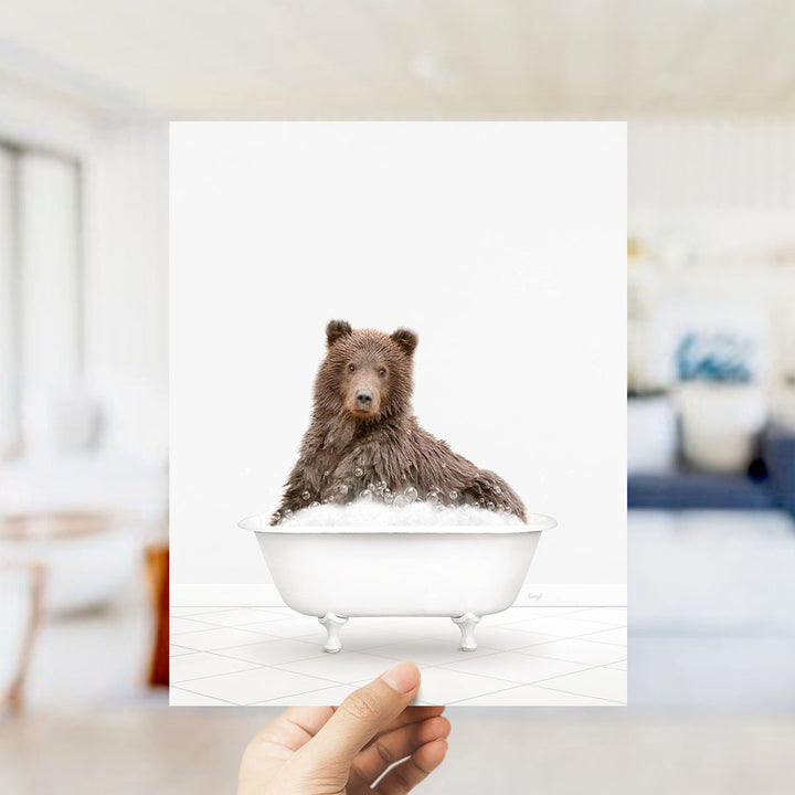 A hand holds up a framed image of a brown bear sitting in a bathtub filled with bubbles.
