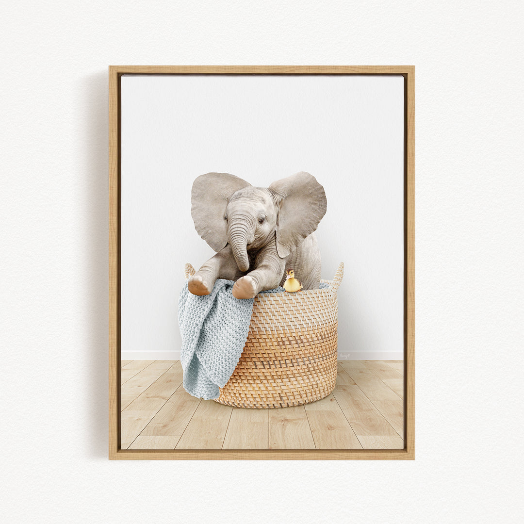 A baby elephant sitting in a woven basket, with a blue blanket and a small yellow toy nearby.