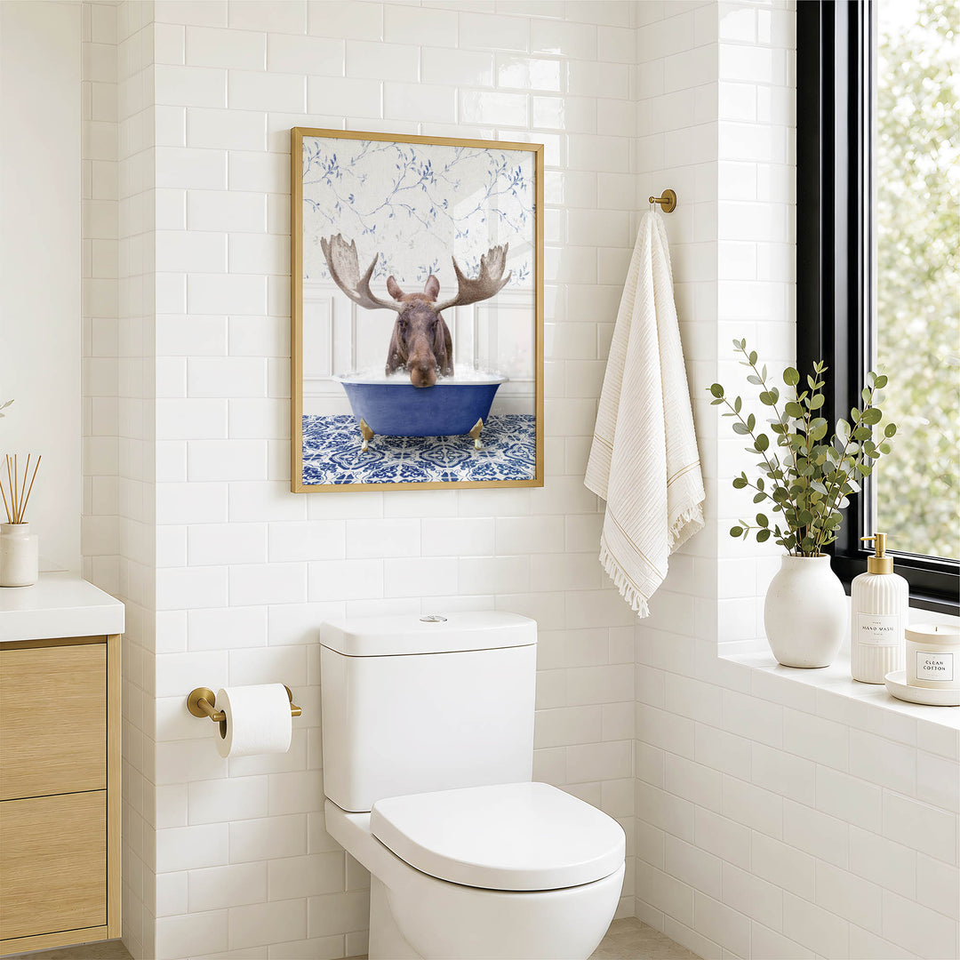 A framed image of a moose in a blue bathtub is displayed on the wall above a white toilet in a bathroom.