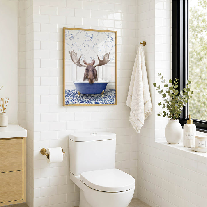 A framed image of a moose in a blue bathtub is displayed on the wall above a white toilet in a bathroom.