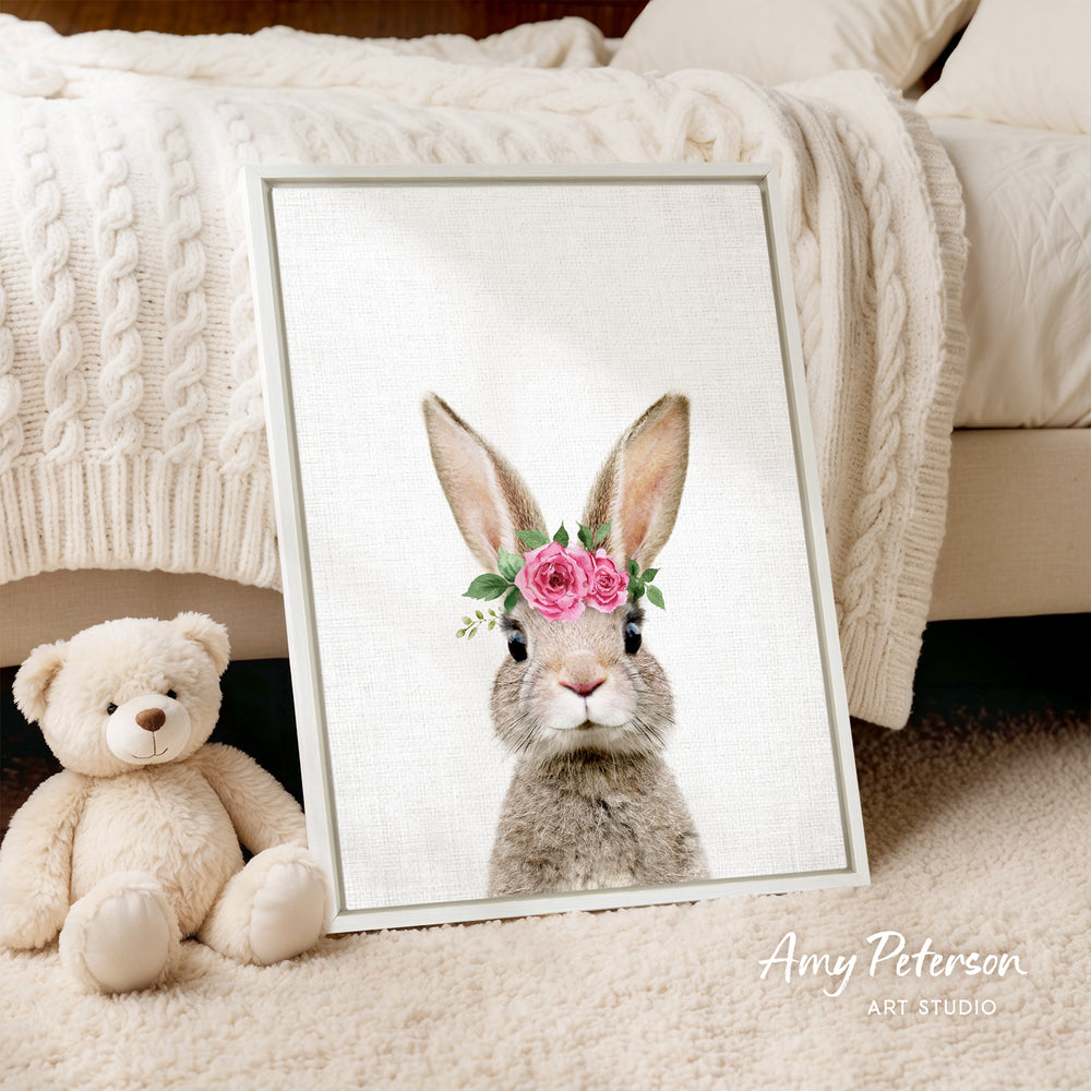 A framed print of a rabbit wearing a flower crown is displayed on a beige carpet, accompanied by a teddy bear.