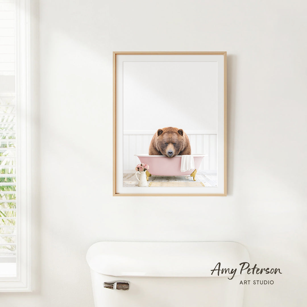 A framed image of a brown bear sitting in a pink bathtub, with a window and toilet visible in the background.
