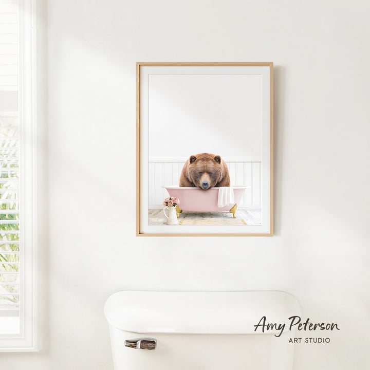 A framed image of a brown bear sitting in a pink bathtub, with a window and toilet visible in the background.