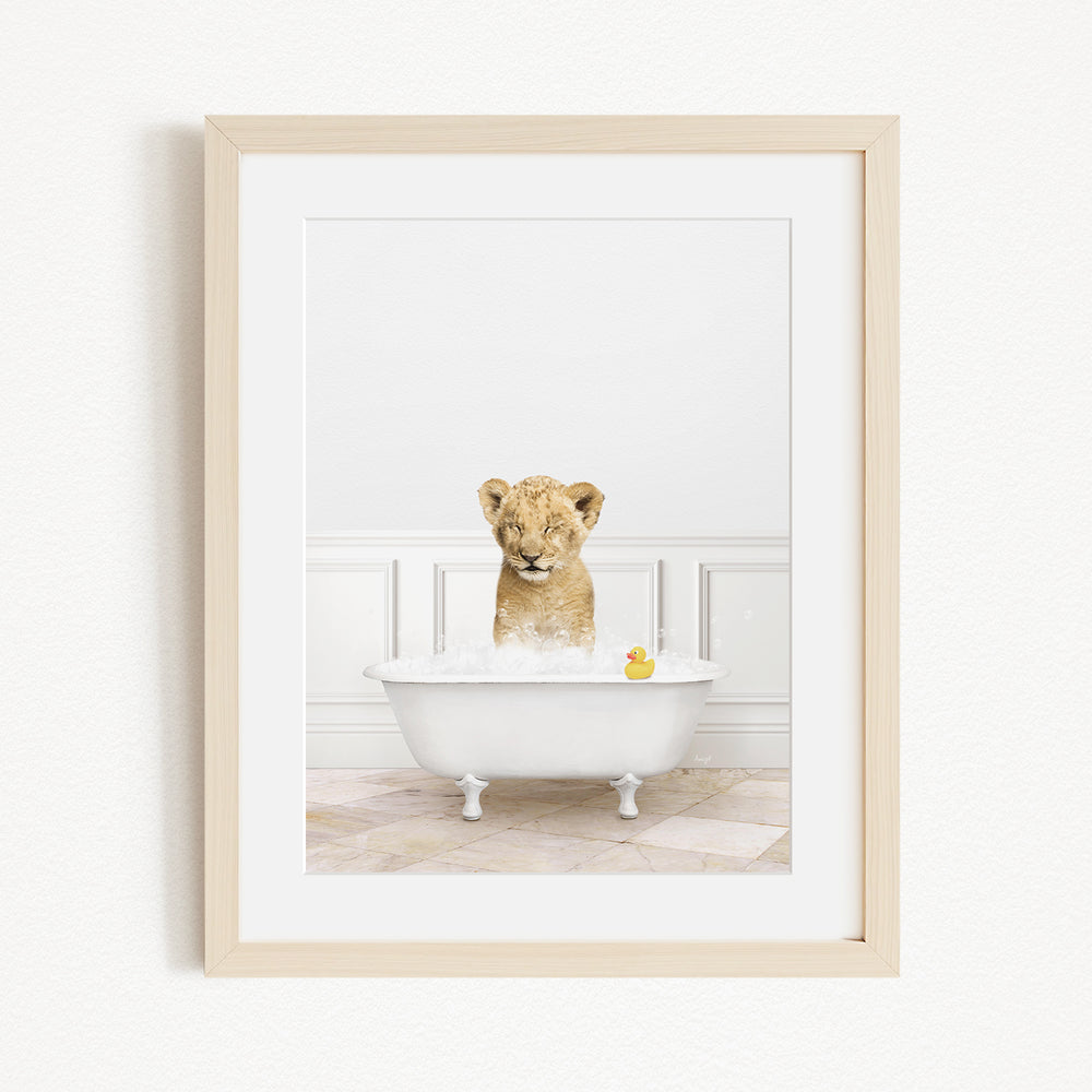 A young lion cub sitting in a white bathtub filled with water and bubbles, with a rubber duck floating in the water.