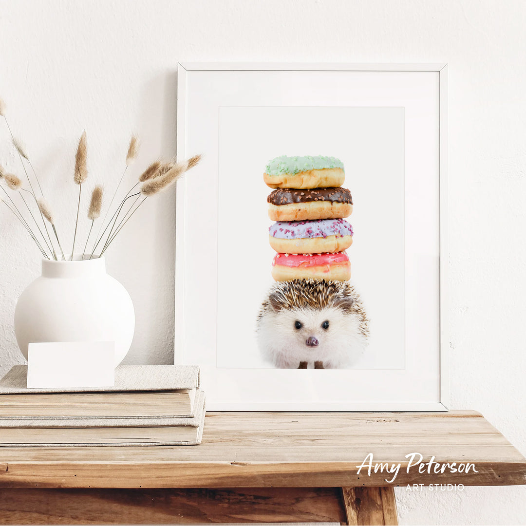 A framed image of a hedgehog sitting on top of a stack of colorful donuts, with a white vase of dried flowers to the left.