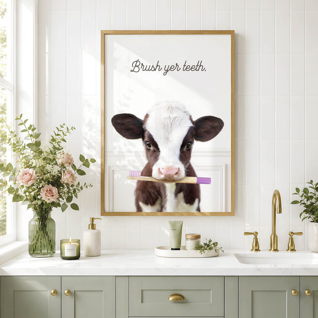 a bathroom scene with a framed photo of a cow holding a toothbrush, a vase of flowers, and a sink with a toothbrush holder.