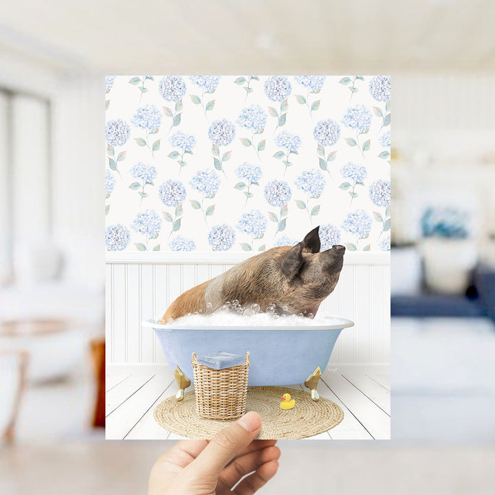 A hand holds up a framed image of a pig sitting in a blue bathtub filled with bubbles, surrounded by a floral patterned wall.