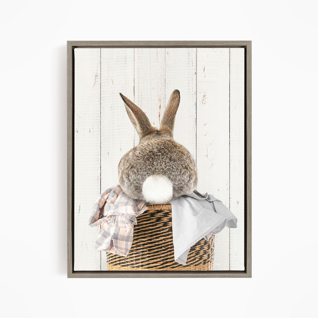 A fluffy gray rabbit sits in a woven basket, with a plaid shirt and a gray blanket placed on top of the basket.