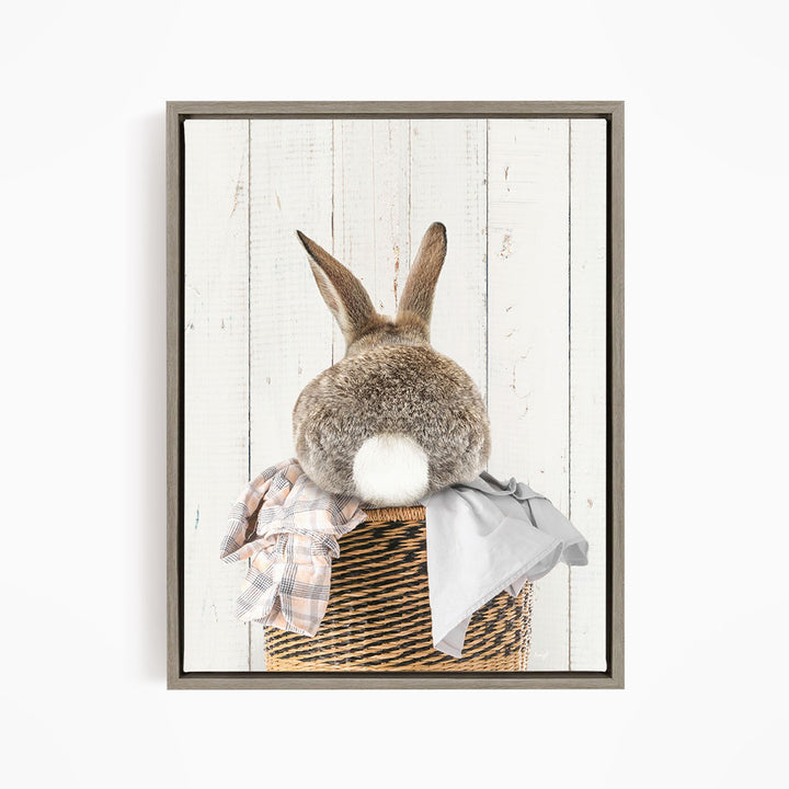 A fluffy gray rabbit sits in a woven basket, with a plaid shirt and a gray blanket placed on top of the basket.