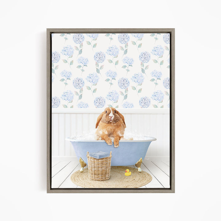 A brown rabbit sitting in a blue bathtub filled with bubbles, surrounded by a wicker basket and rubber duckies, with a floral patterned wall behind it.