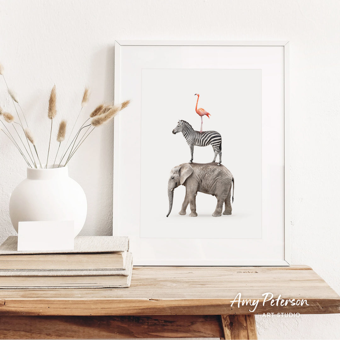 a framed print of a zebra and a flamingo standing on top of an elephant, placed on a wooden shelf next to a white vase with dried flowers.