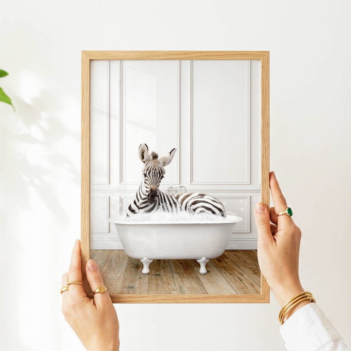 A person's hands are holding a picture frame with a zebra resting in a white bathtub, creating a whimsical and playful scene.