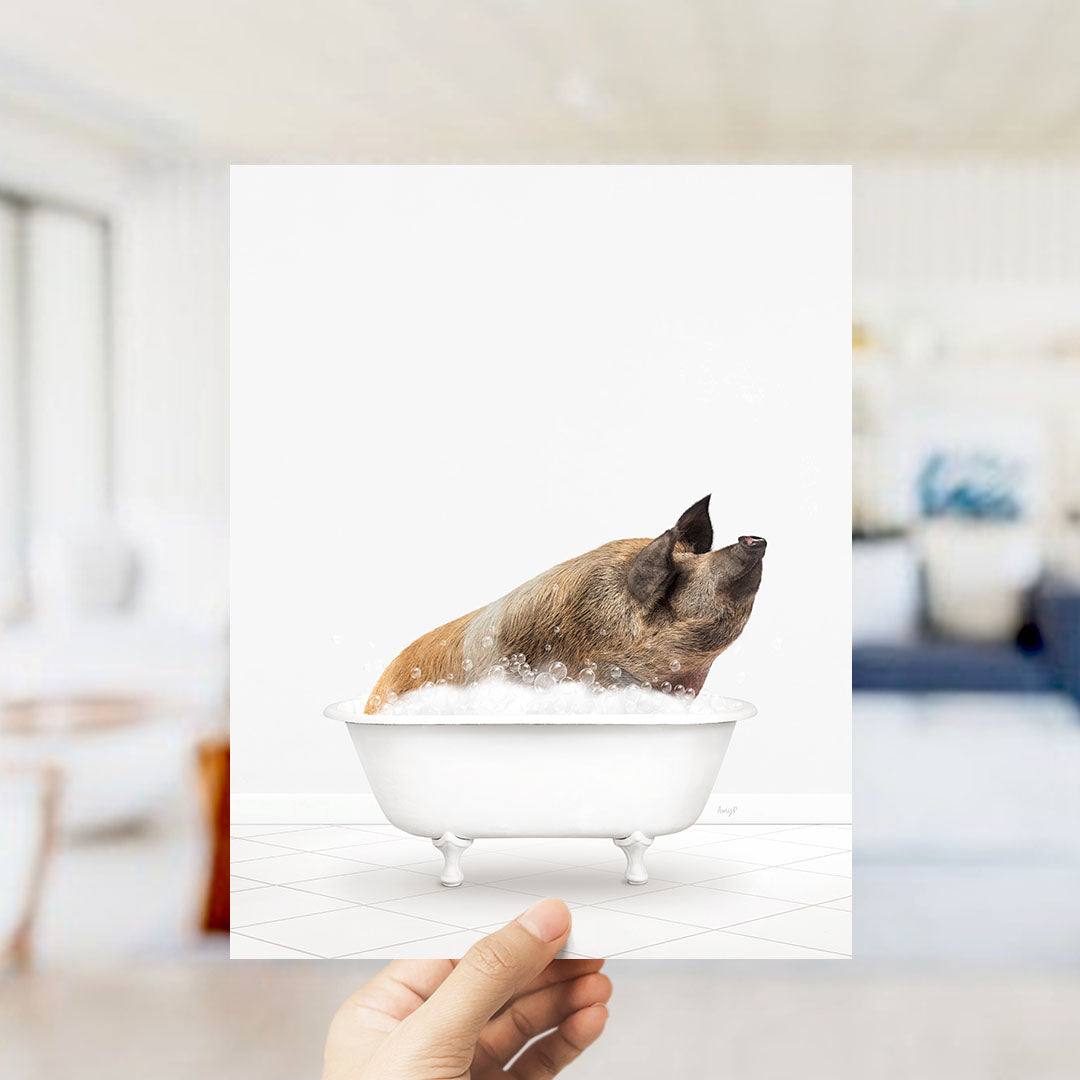 A hand holds up a framed image of a pig sitting in a bathtub filled with bubbles.