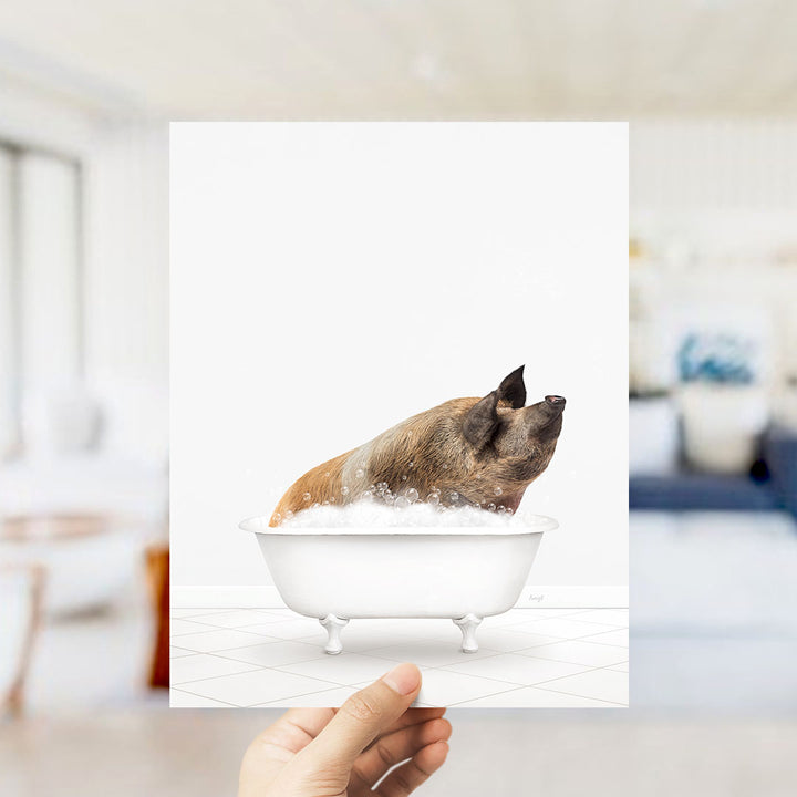 A hand holds up a framed image of a pig sitting in a bathtub filled with bubbles.