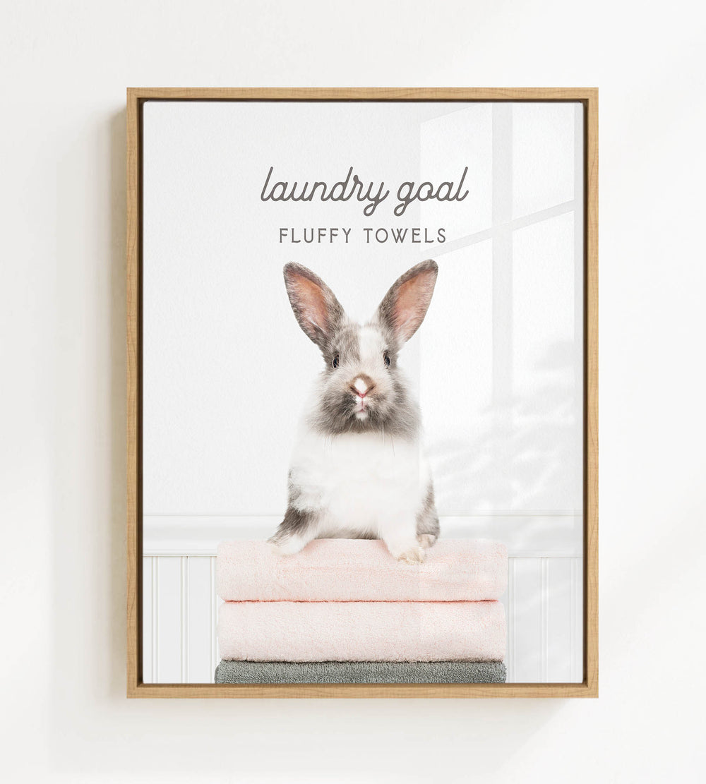 A fluffy white and gray rabbit sitting on a stack of pink towels, with the text "laundry goal FLUFFY TOWELS" displayed above the rabbit.