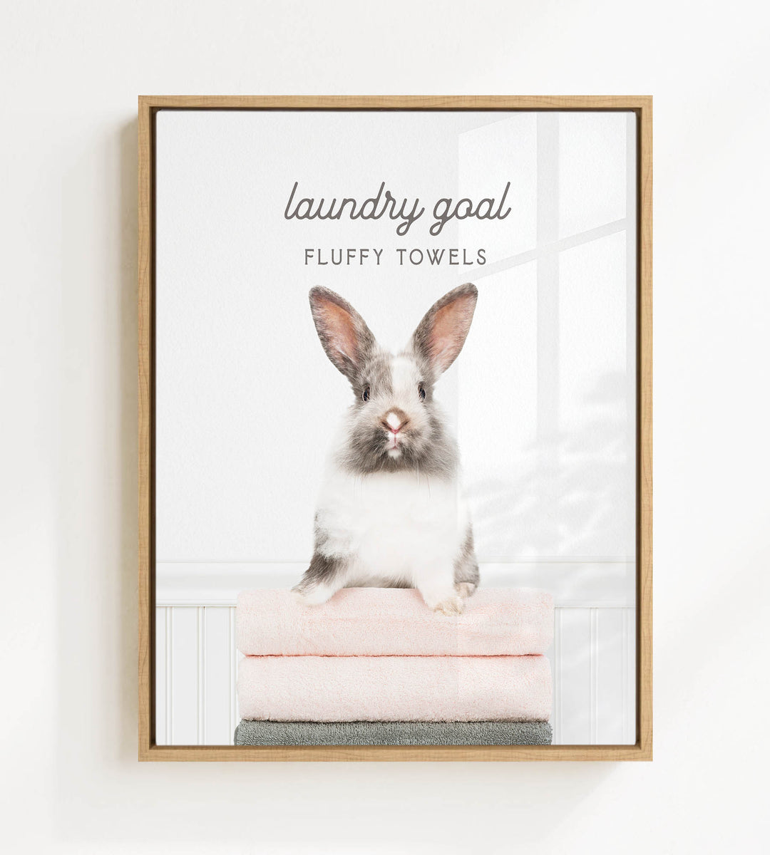 A fluffy white and gray rabbit sitting on a stack of pink towels, with the text "laundry goal FLUFFY TOWELS" displayed above the rabbit.