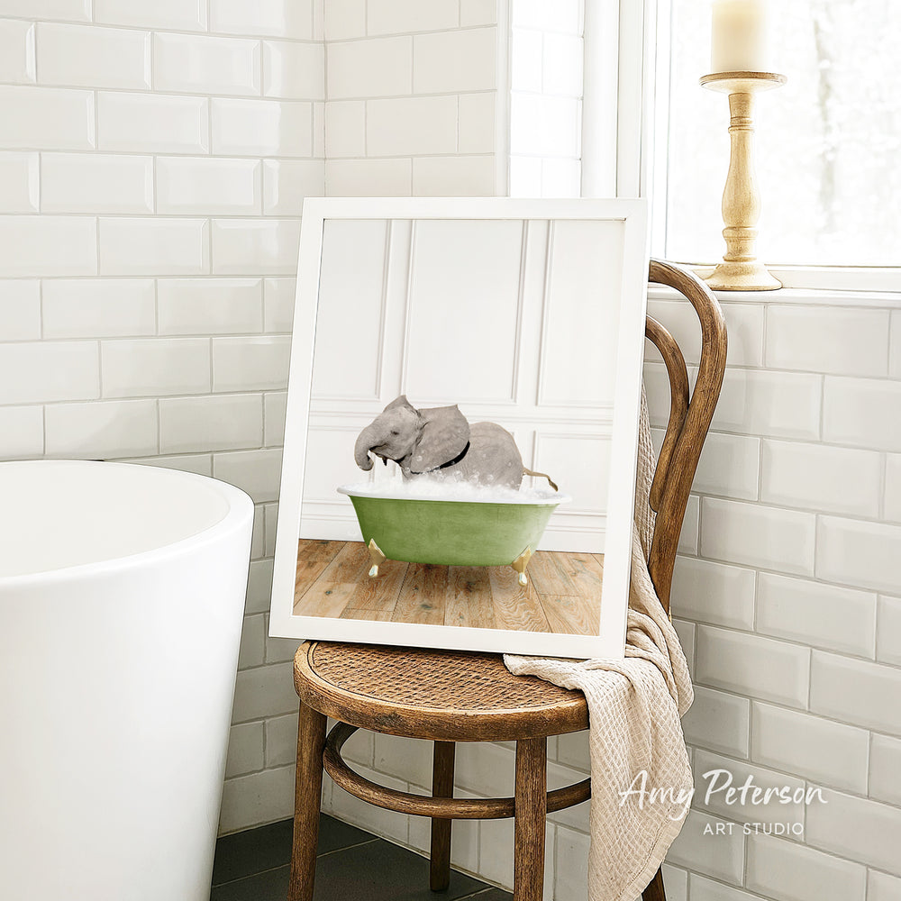 A framed image of a gray elephant sitting in a green bathtub, with a wooden chair and a white tiled wall in the background.
