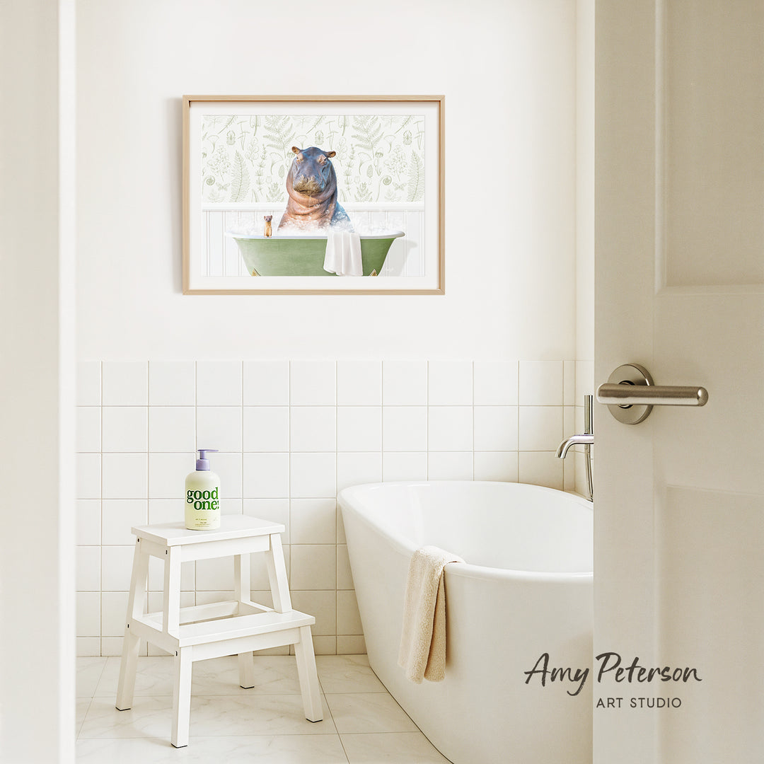 A framed painting of a hippopotamus sitting in a bathtub is hanging on a white tiled wall above a white bathtub in a bathroom.