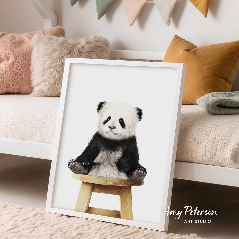 A framed print of a panda sitting on a wooden stool is displayed in a cozy bedroom setting with a white bed and colorful pillows.