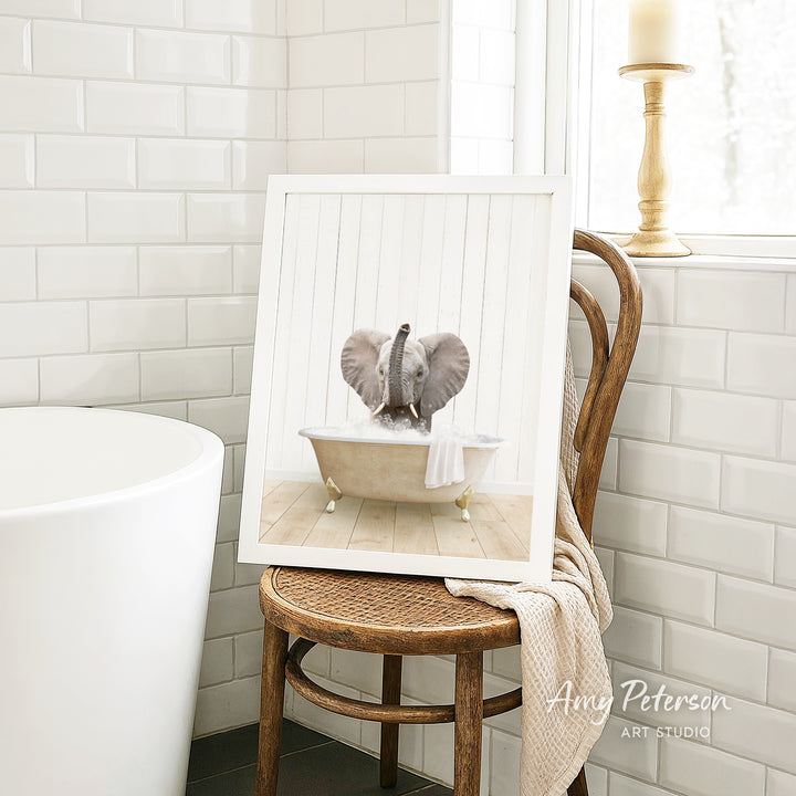 A framed image of a baby elephant sitting in a bathtub, with a towel draped over the side of the tub.