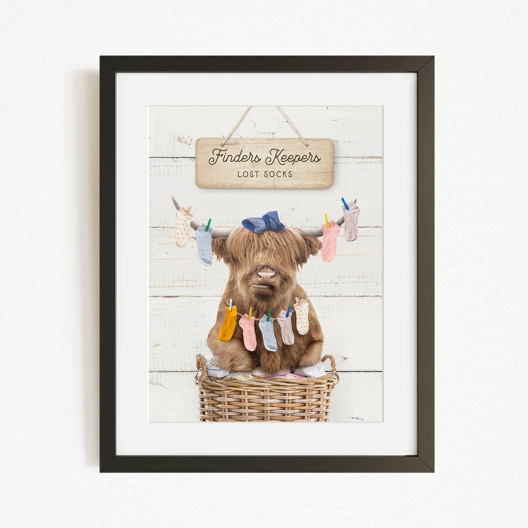 A framed image depicting a brown cow sitting in a wicker basket, with socks hanging from its horns.