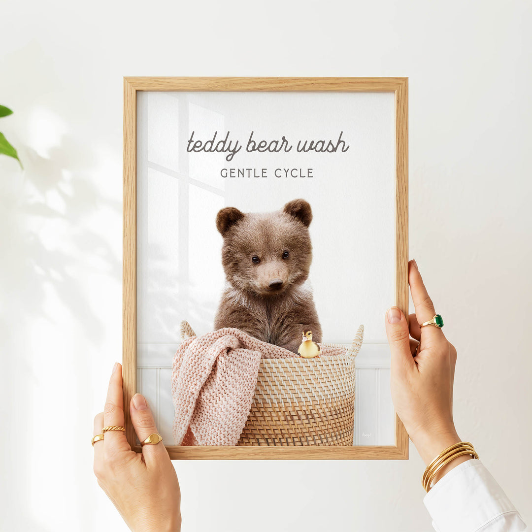 A person's hands are holding a framed picture of a teddy bear in a basket, with the text "teddy bear wash" and "GENTLE CYCLE" written above the bear.