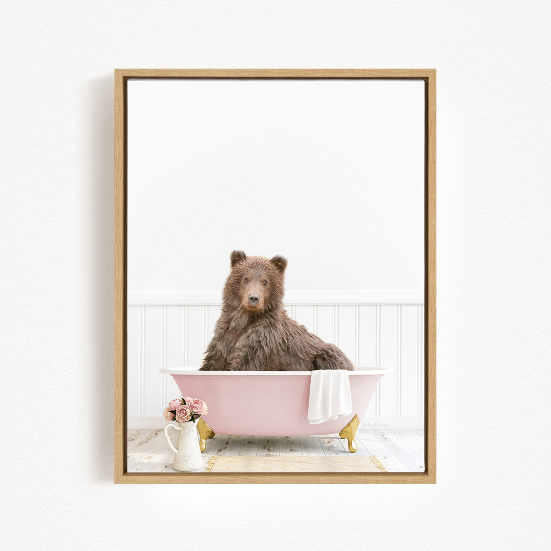 A brown bear sitting in a pink bathtub, with a vase of flowers and a towel nearby.