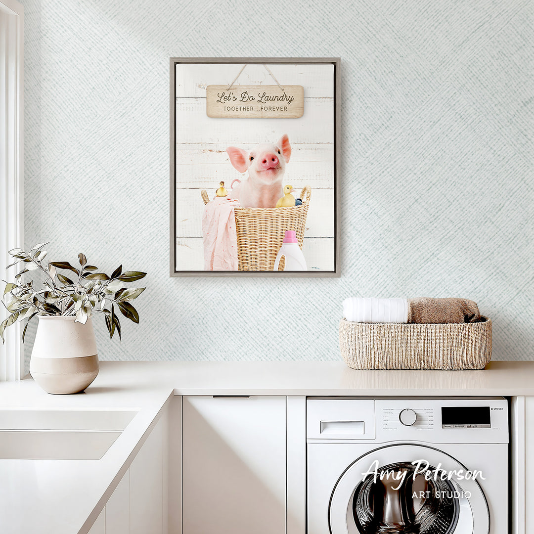 a framed picture of a pig sitting in a basket, with a sign above it that says "Let's Do Laundry Together Forever". The scene is set in a kitchen or laundry room, with a white sink, a white washing machine, and a potted plant nearby.