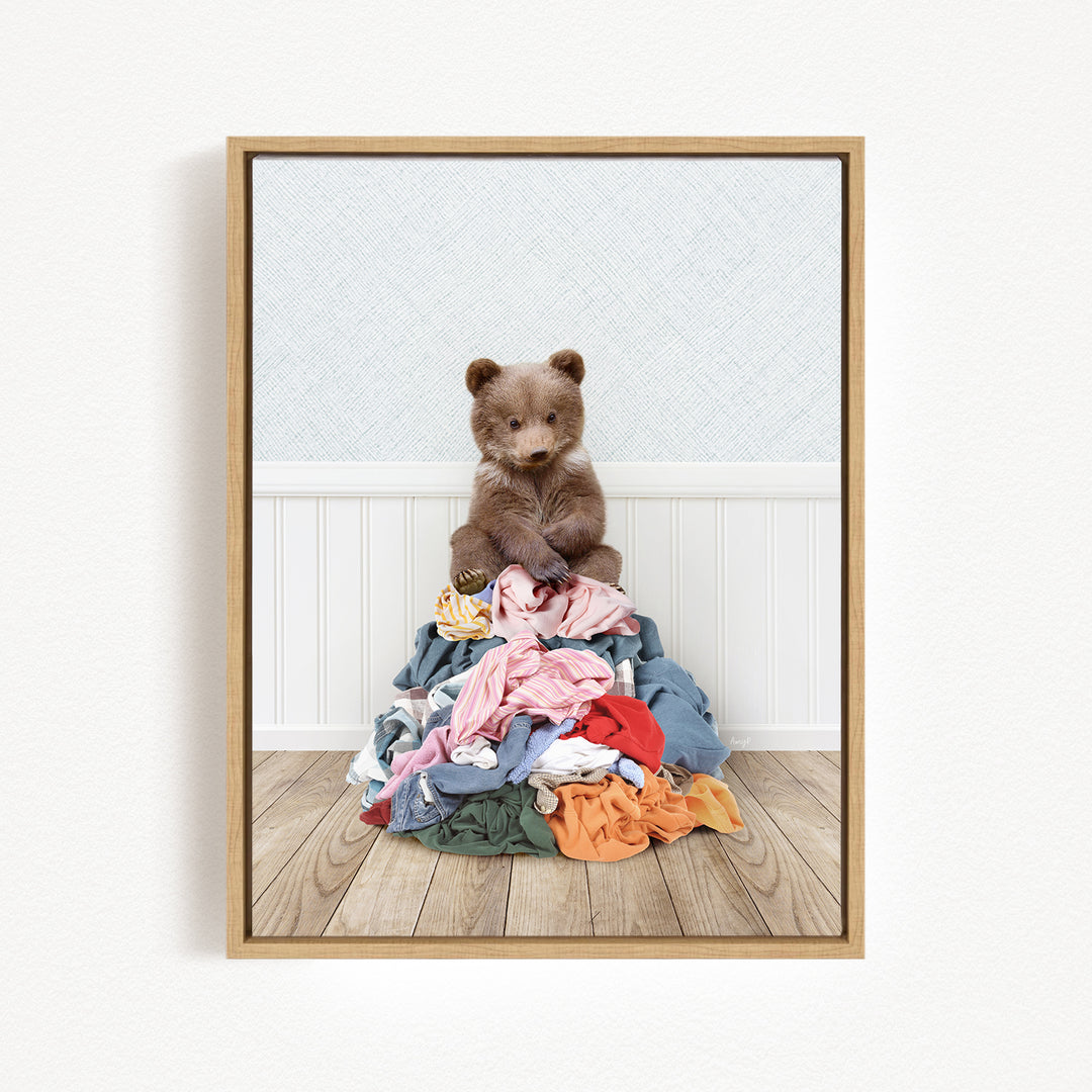 A teddy bear sits atop a pile of clothes on a wooden floor.