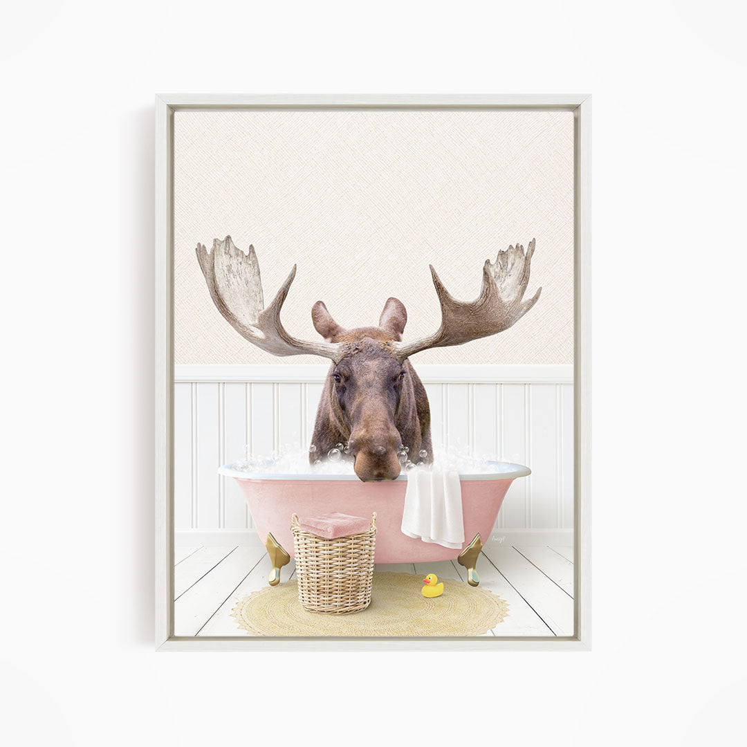 A moose with large antlers is sitting in a pink bathtub filled with water, surrounded by a yellow rubber duck and a wicker basket.