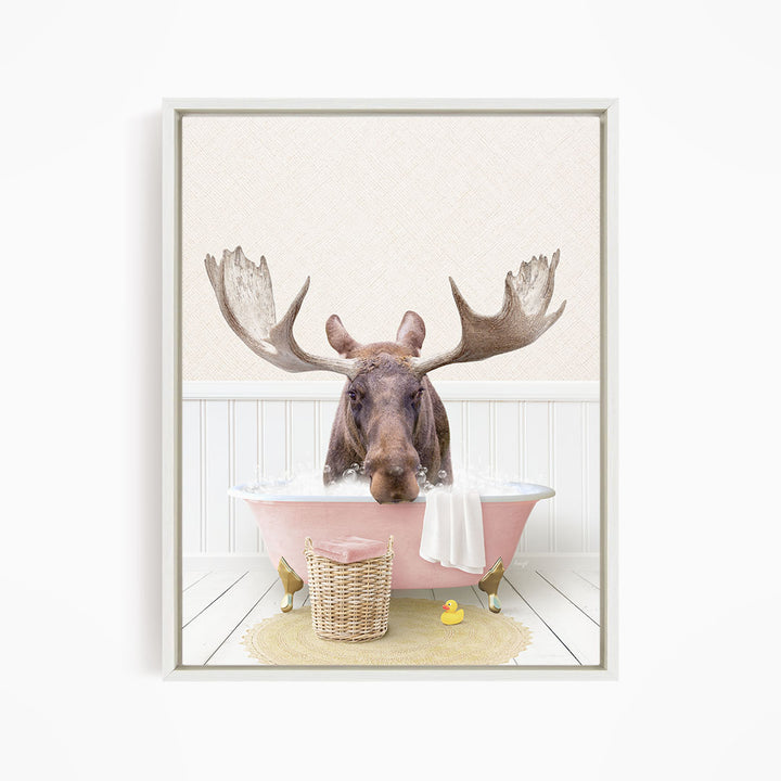 A moose with large antlers is sitting in a pink bathtub filled with water, surrounded by a yellow rubber duck and a wicker basket.