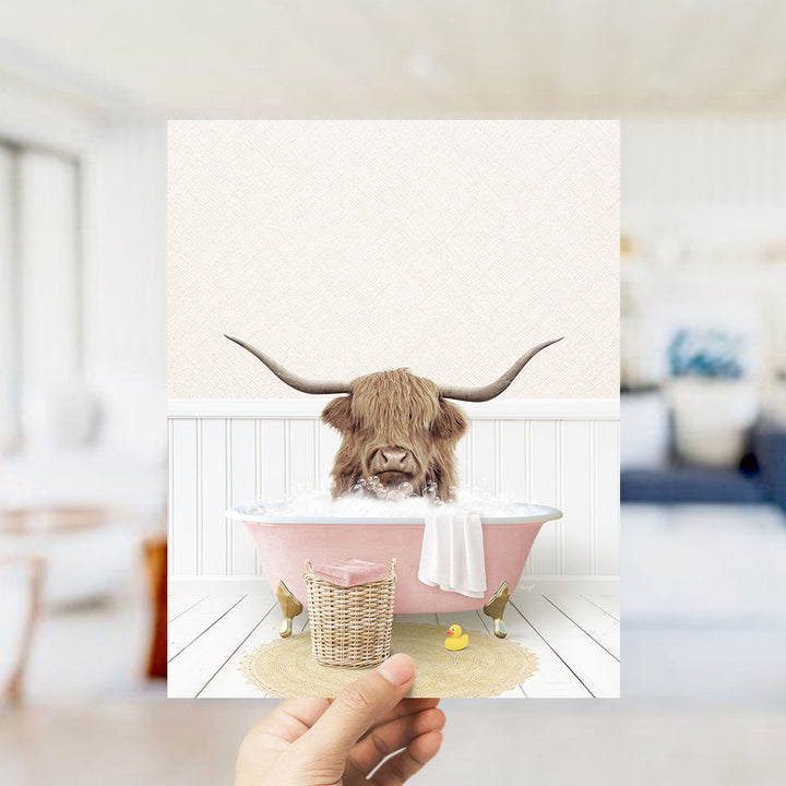 A hand holds up a framed image depicting a Highland cow with large horns sitting in a pink bathtub, surrounded by a yellow rubber duck and a white towel.