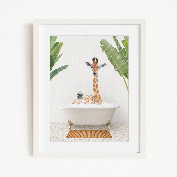 A giraffe sitting in a white bathtub filled with bubbles, surrounded by green leaves and a wooden mat on the floor.